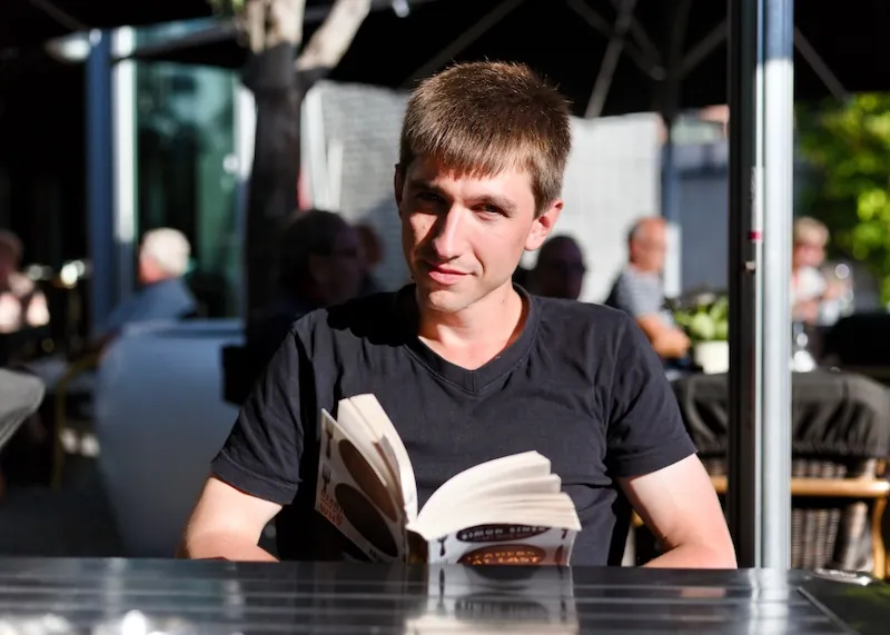 Kristjan Vingel sitting in a outside cafe holding a open book and looking into a camera